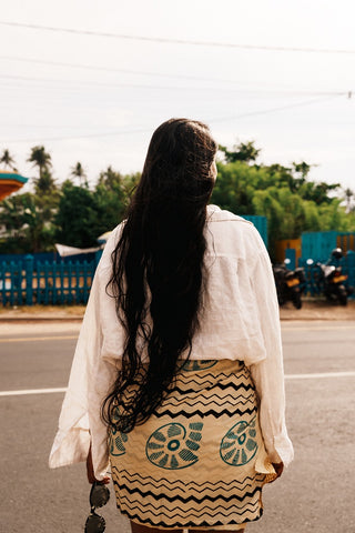 Person wearing a white shirt and patterned skirt on a street with greenery in the background